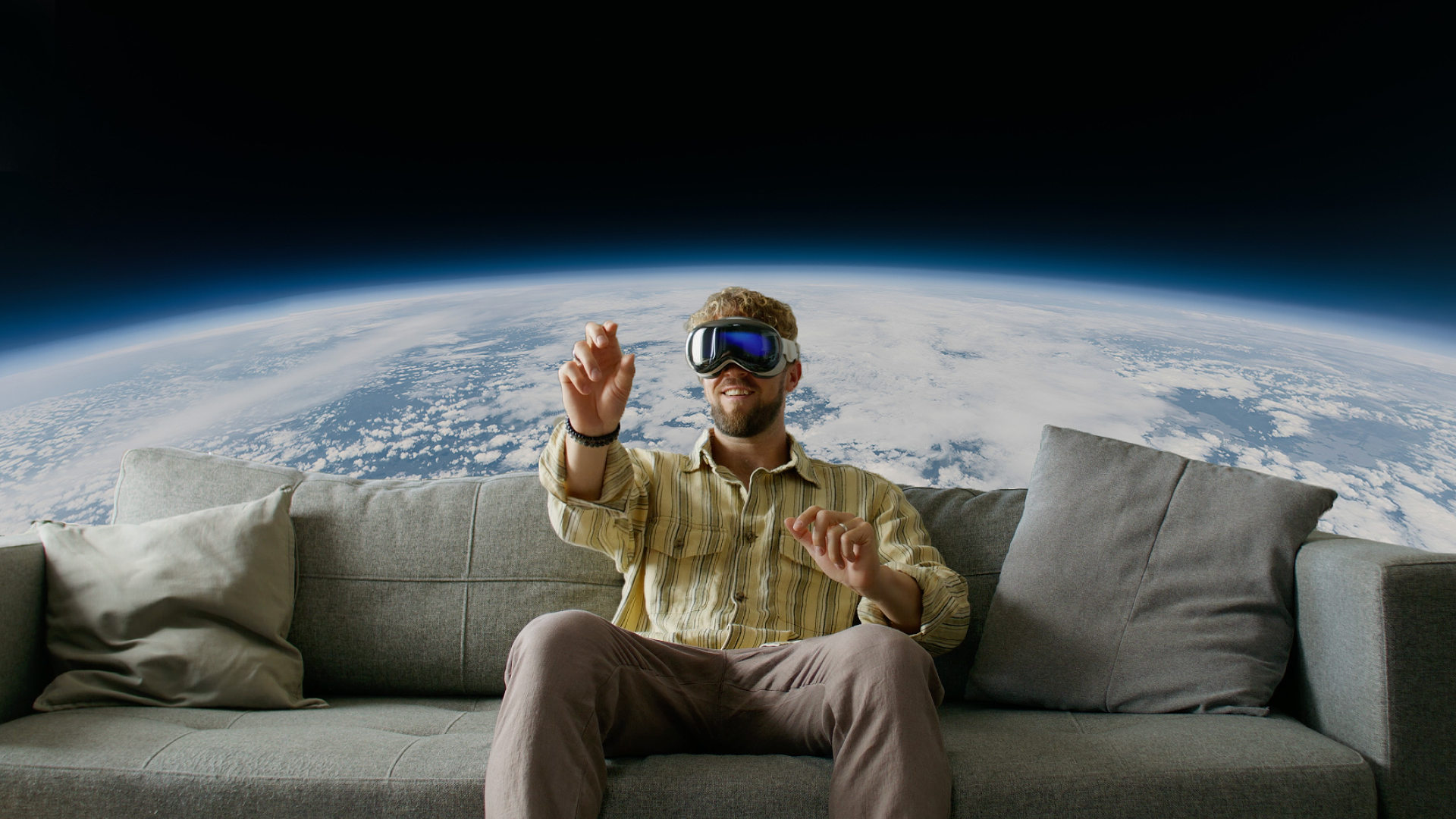 A man sits on a gray couch wearing an Apple Vision Pro, reaching up with both hands as if interacting with something in the air, with a massive curved view of Earth's atmosphere and cloud-covered surface filling the background behind the couch