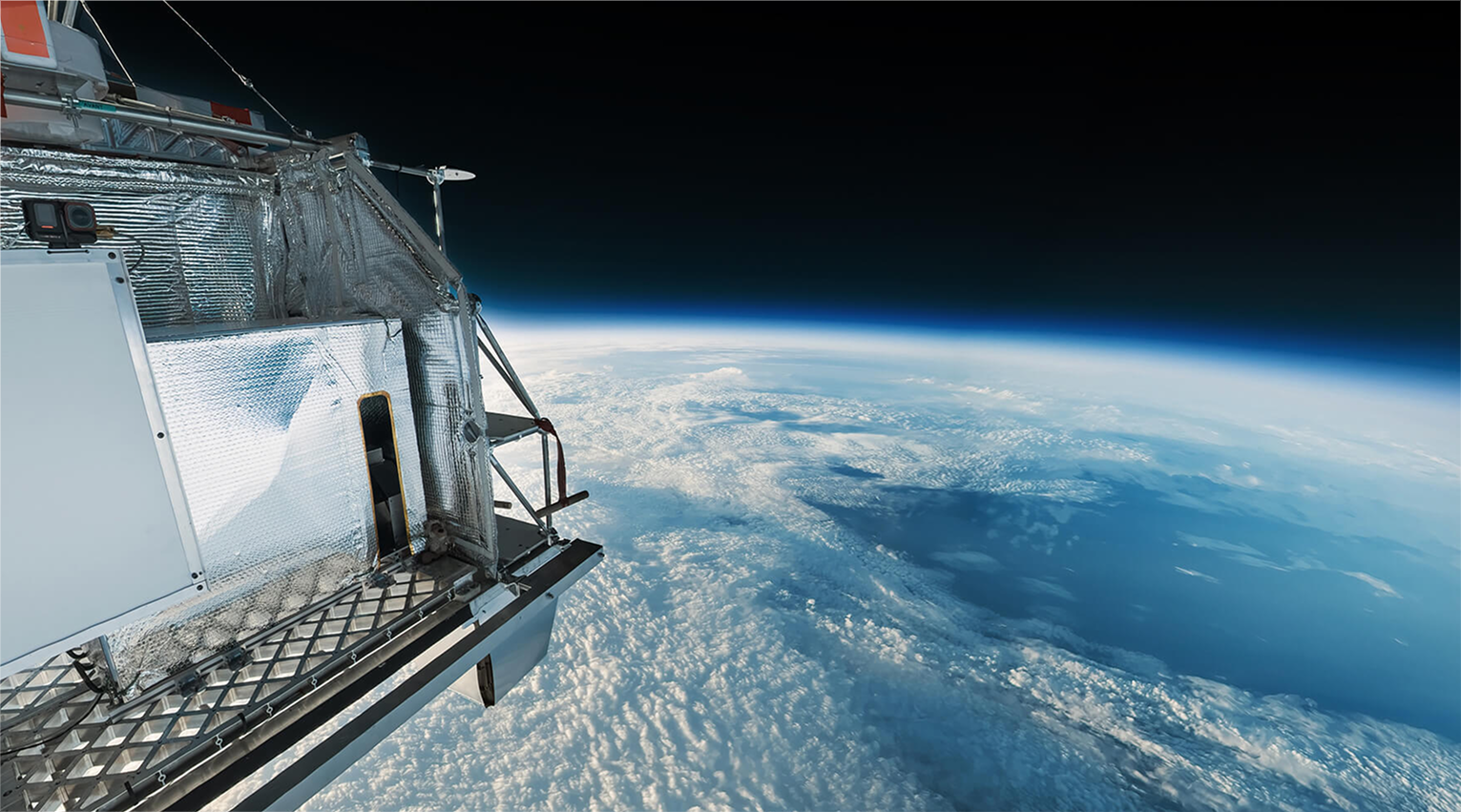 View from a stratospheric balloon payload looking out over Earth's curved horizon, with a metallic/foil-wrapped equipment housing visible in the foreground and dense white clouds below
